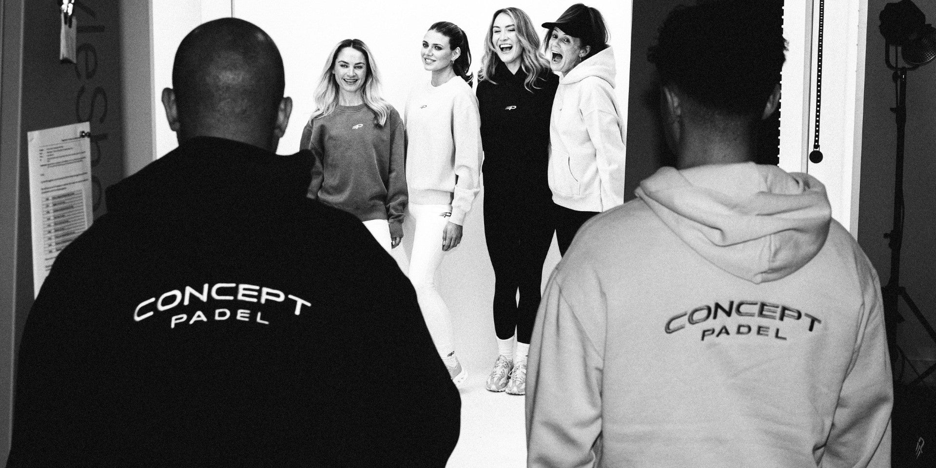 Black‑and‑white behind‑the‑scenes shot of two people in Concept Padel hoodies watching four people pose in front of a studio backdrop with lighting equipment
