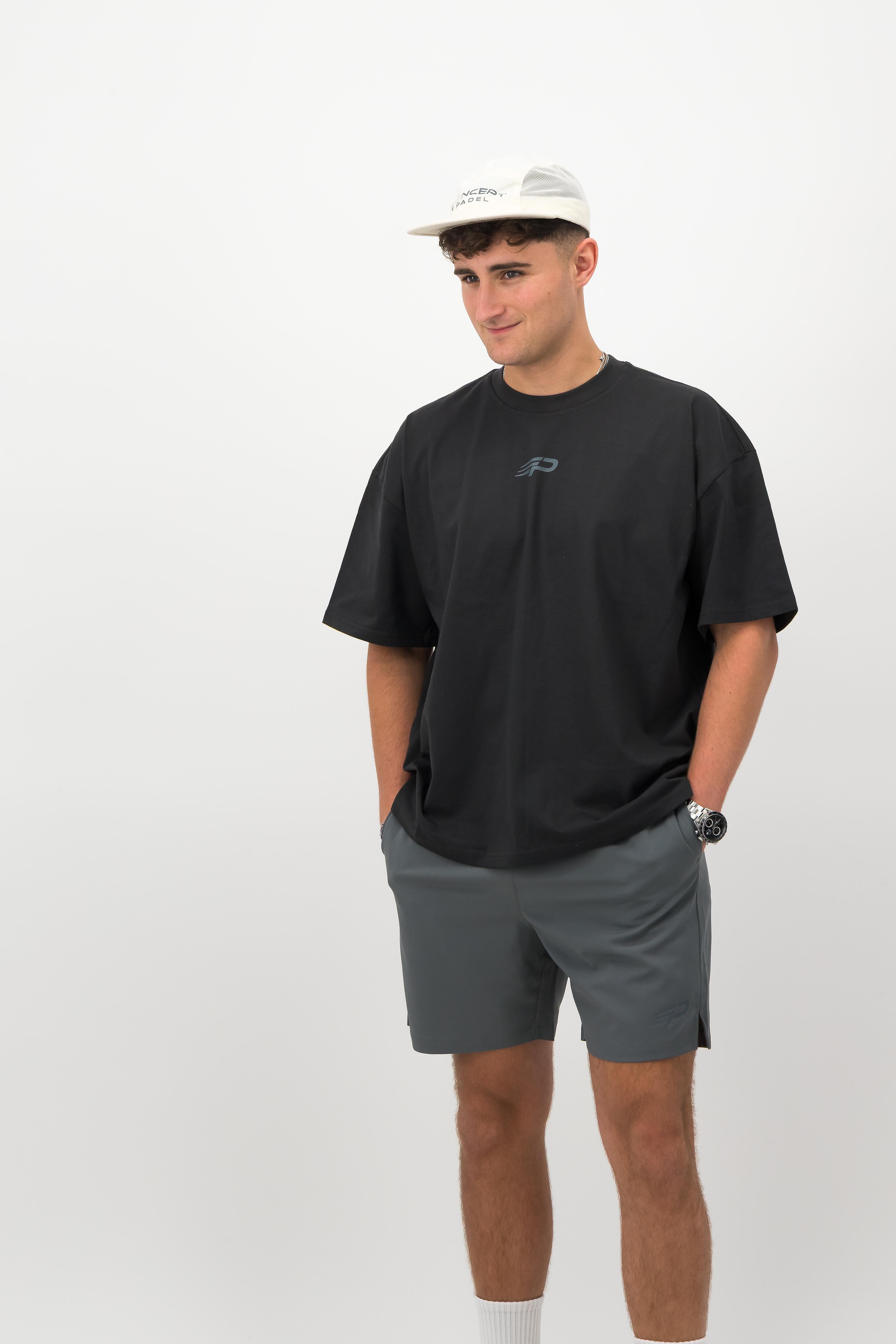 Man wearing ecru Concept Padel performance cap with black oversized T‑shirt and grey shorts front view