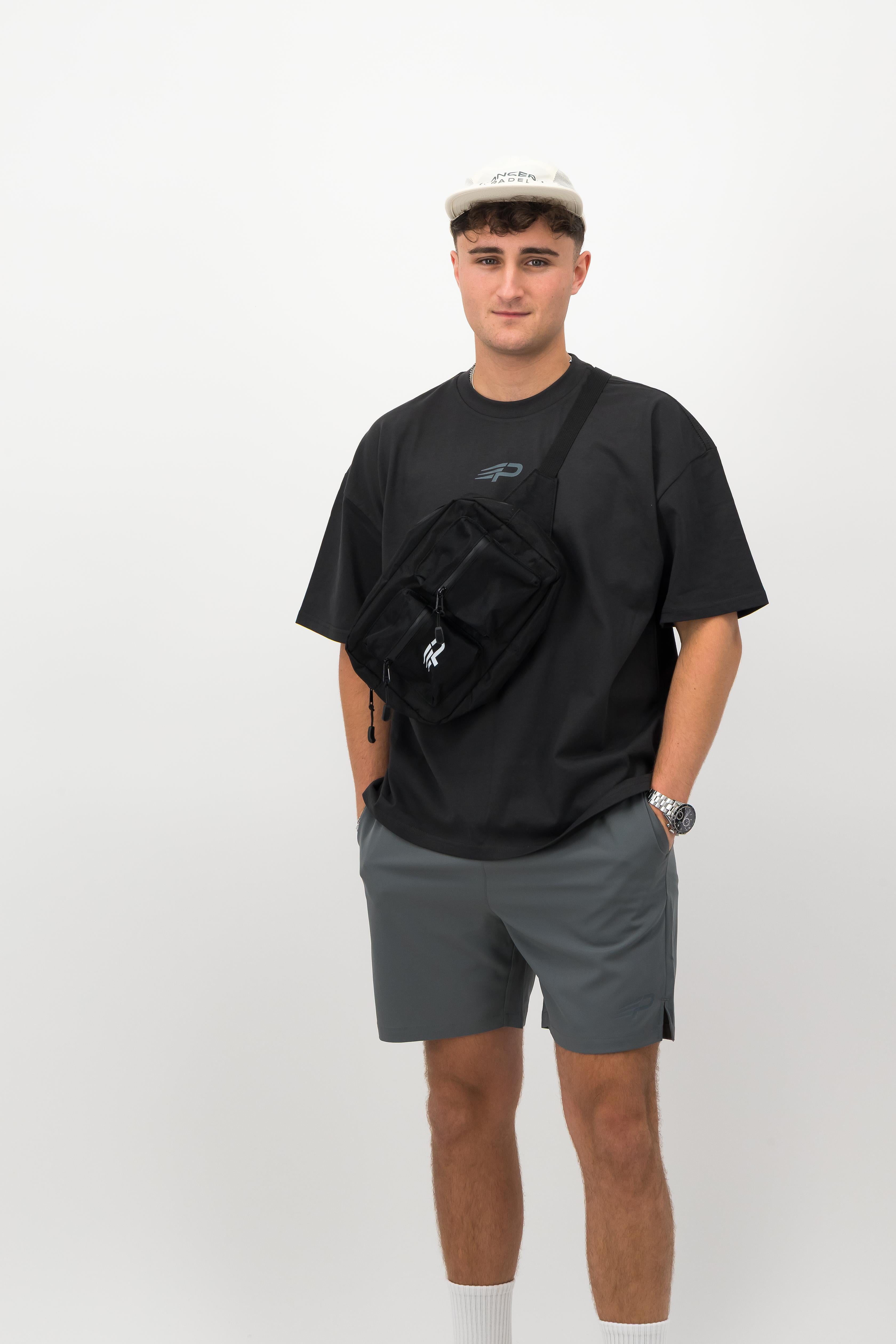 Man wearing ecru performance cap, black T‑shirt, grey shorts and crossbody bag