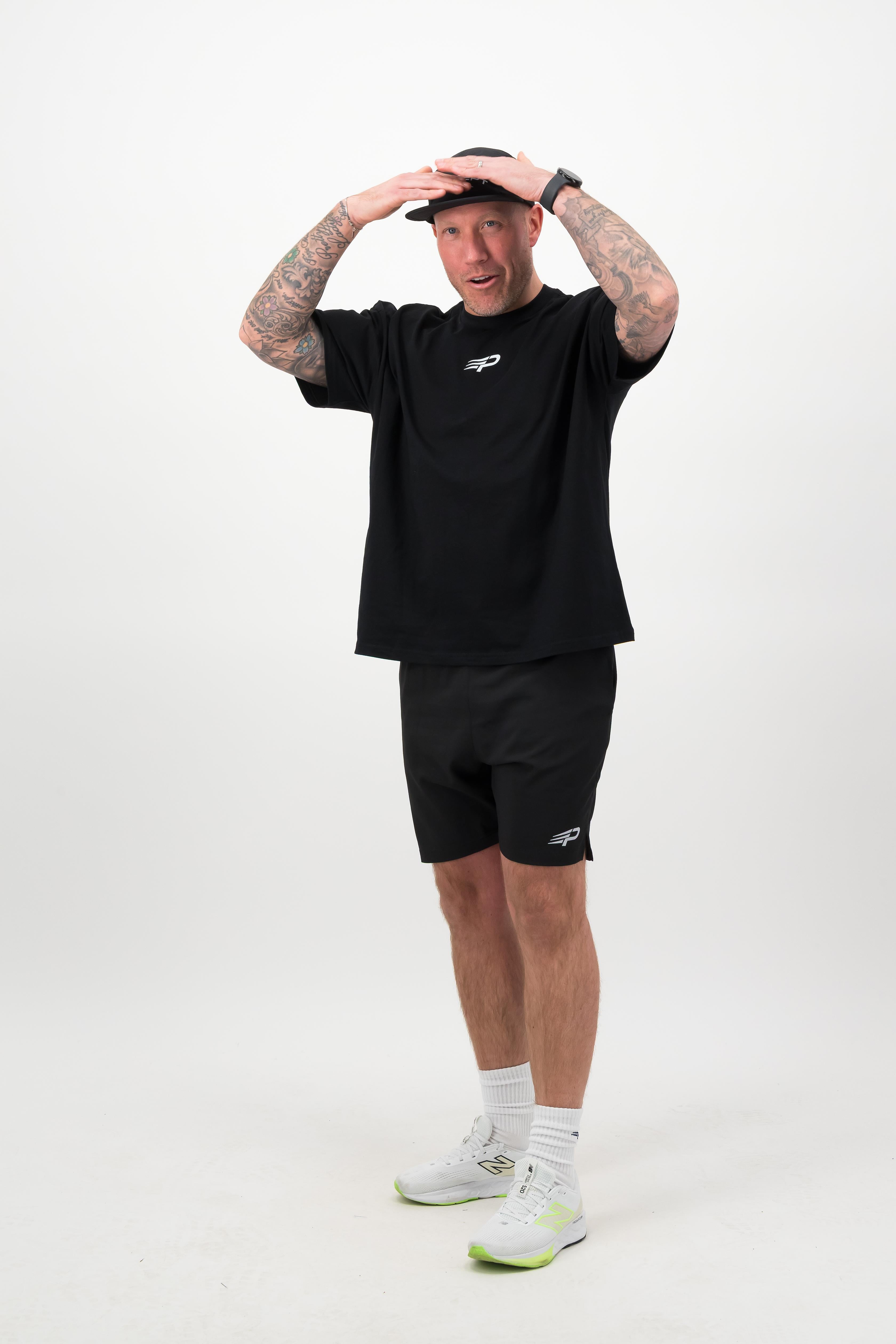 Person wearing a black T‑shirt, black shorts and a black cap with hands on head against a white background