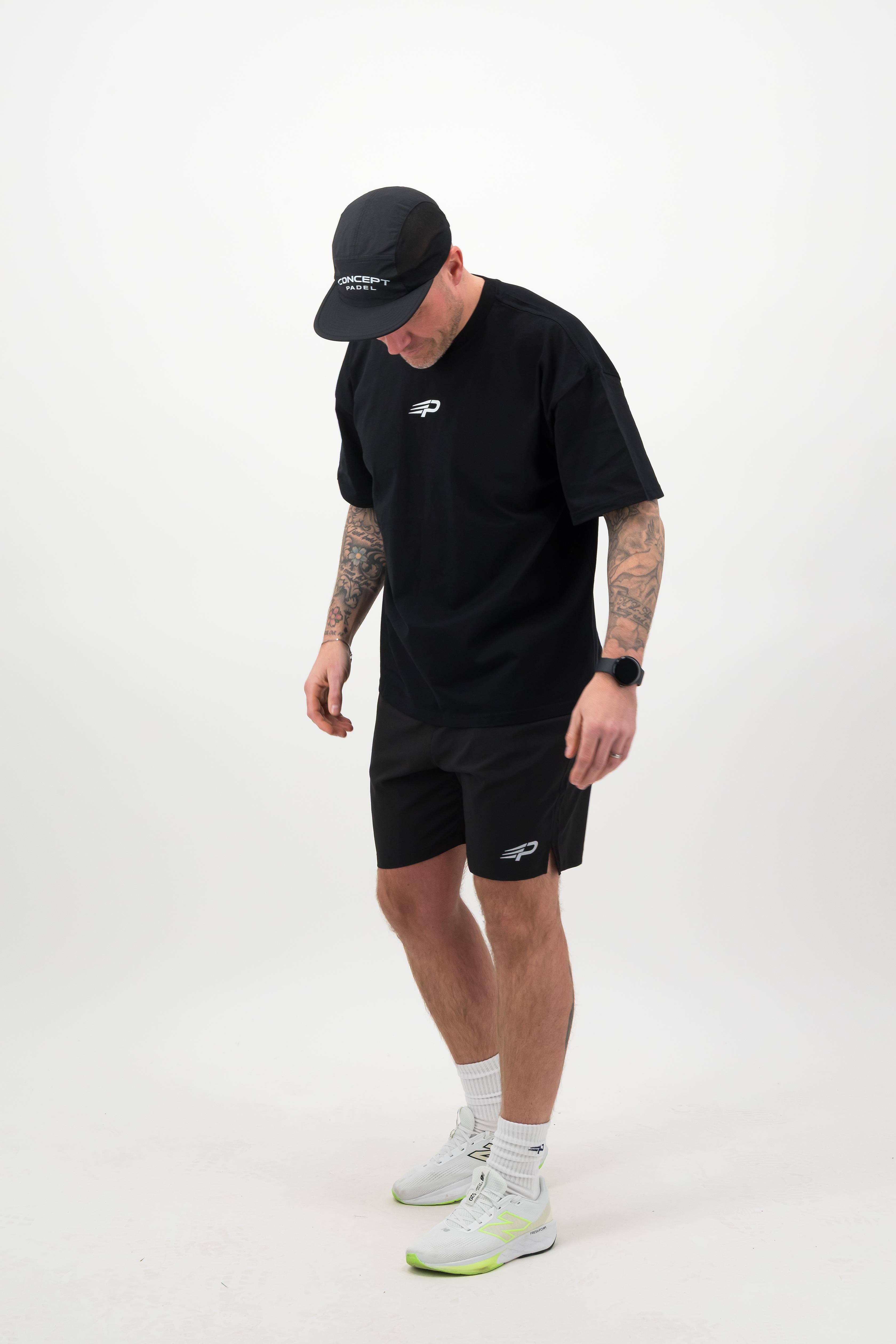 Person wearing a black T‑shirt, black shorts and a black cap looking down against a white background