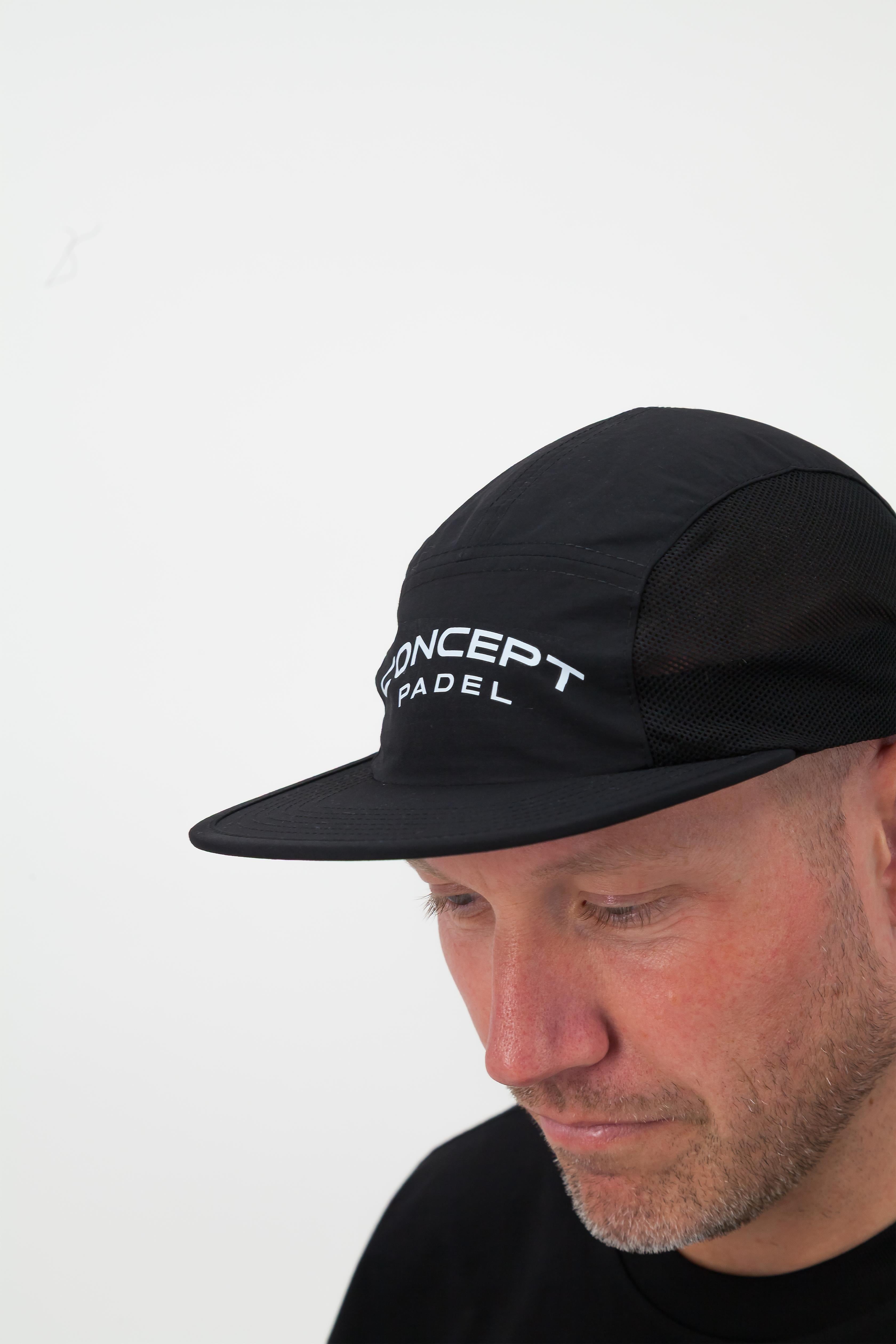 Person wearing a black T‑shirt, black shorts and a black cap looking downward against a white background
