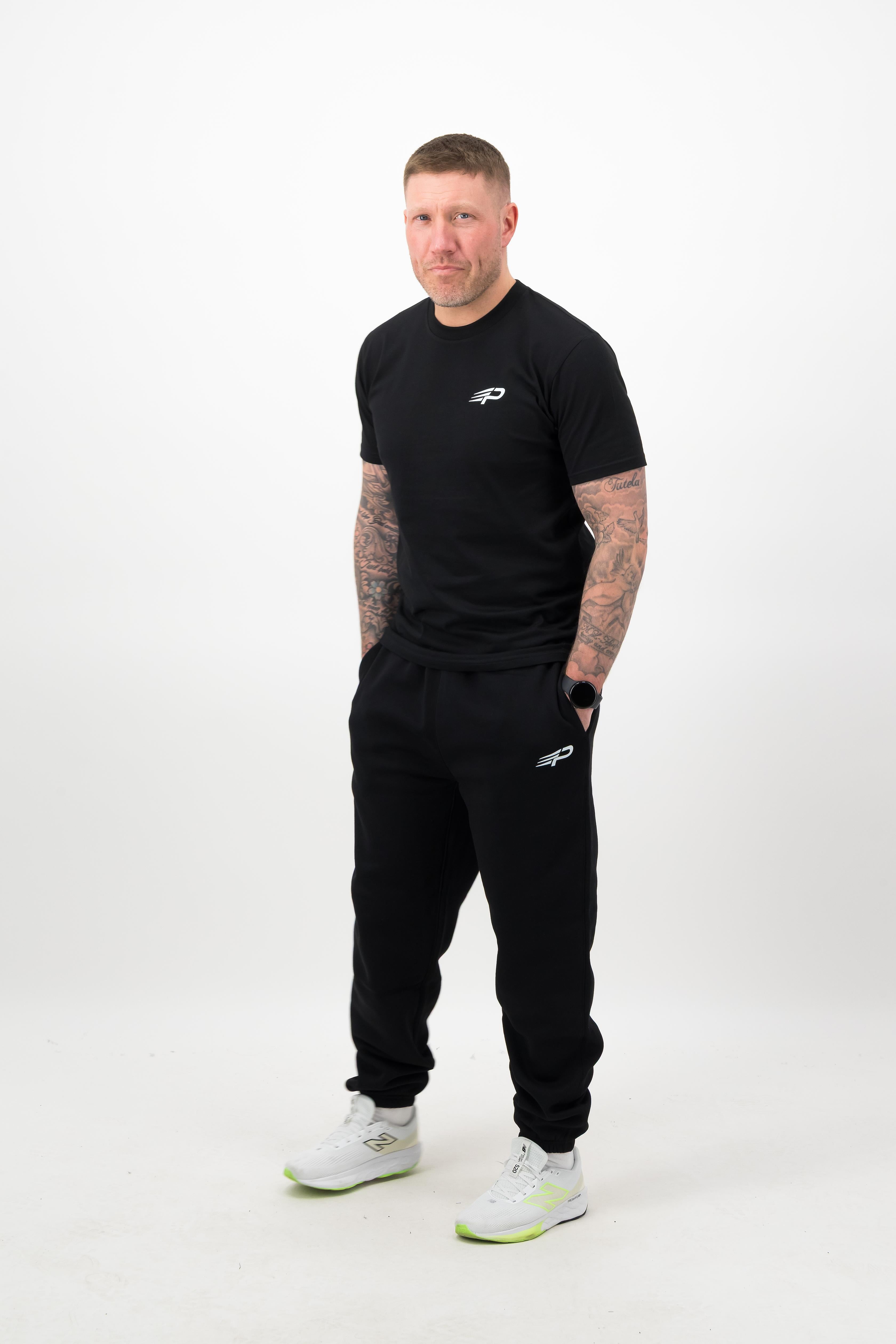 Person wearing a Concept Padel black rebound tee standing facing forward, styled with black joggers and trainers against a white background