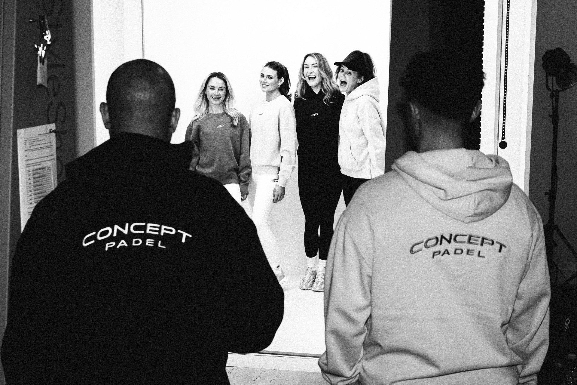 Black‑and‑white behind‑the‑scenes shot of two people in Concept Padel hoodies watching four people pose in front of a studio backdrop with lighting equipment