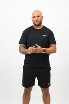Person wearing a Concept Padel black performance tee standing facing forward, styled with dark shorts and trainers against a white background