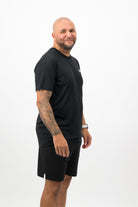 Person wearing a Concept Padel black performance tee standing side on, paired with dark shorts and trainers against a white background