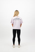Woman wearing a Concept Padel orchid court tee standing with back to camera showing the relaxed silhouette, styled with black shorts and trainers against a white background