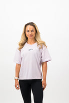 Woman wearing a Concept Padel orchid court tee standing in a relaxed pose with weight shifted to one leg, paired with black shorts and trainers against a white background