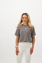 Woman wearing a Concept Padel grey faded crop tee standing facing forward, styled with white leggings and trainers against a white background