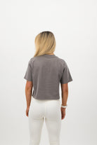 Woman wearing a Concept Padel grey faded crop tee standing with back to camera showing the clean back silhouette, styled with white leggings and trainers against a white background