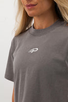 Close‑up of the embroidered Concept Padel logo on the grey faded crop tee worn by a woman against a white background