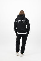 Woman wearing a Concept Padel black serve hoodie standing with back to camera showing the back arch logo, styled with matching black serve sweatpants and trainers against a white background