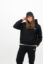 Woman wearing a Concept Padel black serve hoodie standing facing forward, styled with matching black serve sweatpants and trainers against a white background