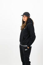 Woman wearing a Concept Padel black serve hoodie standing side on, paired with matching black serve sweatpants and trainers against a white background
