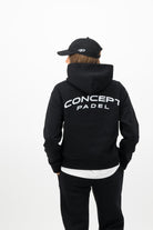Woman wearing a Concept Padel black serve hoodie standing with back to camera showing the back arch logo, styled with matching black serve sweatpants and trainers against a white background