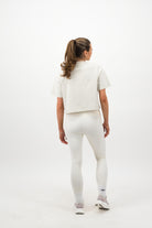 Woman wearing a Concept Padel bone faded crop tee standing with back to camera showing the clean back silhouette, styled with white leggings and trainers against a white background