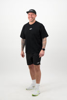 Person wearing a black T‑shirt, black shorts and a black cap standing and smiling against a white background