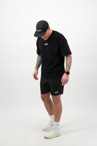 Person wearing a black T‑shirt, black shorts and a black cap looking down against a white background