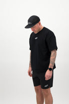 Person wearing a black T‑shirt, black shorts and a black cap looking downward against a white background