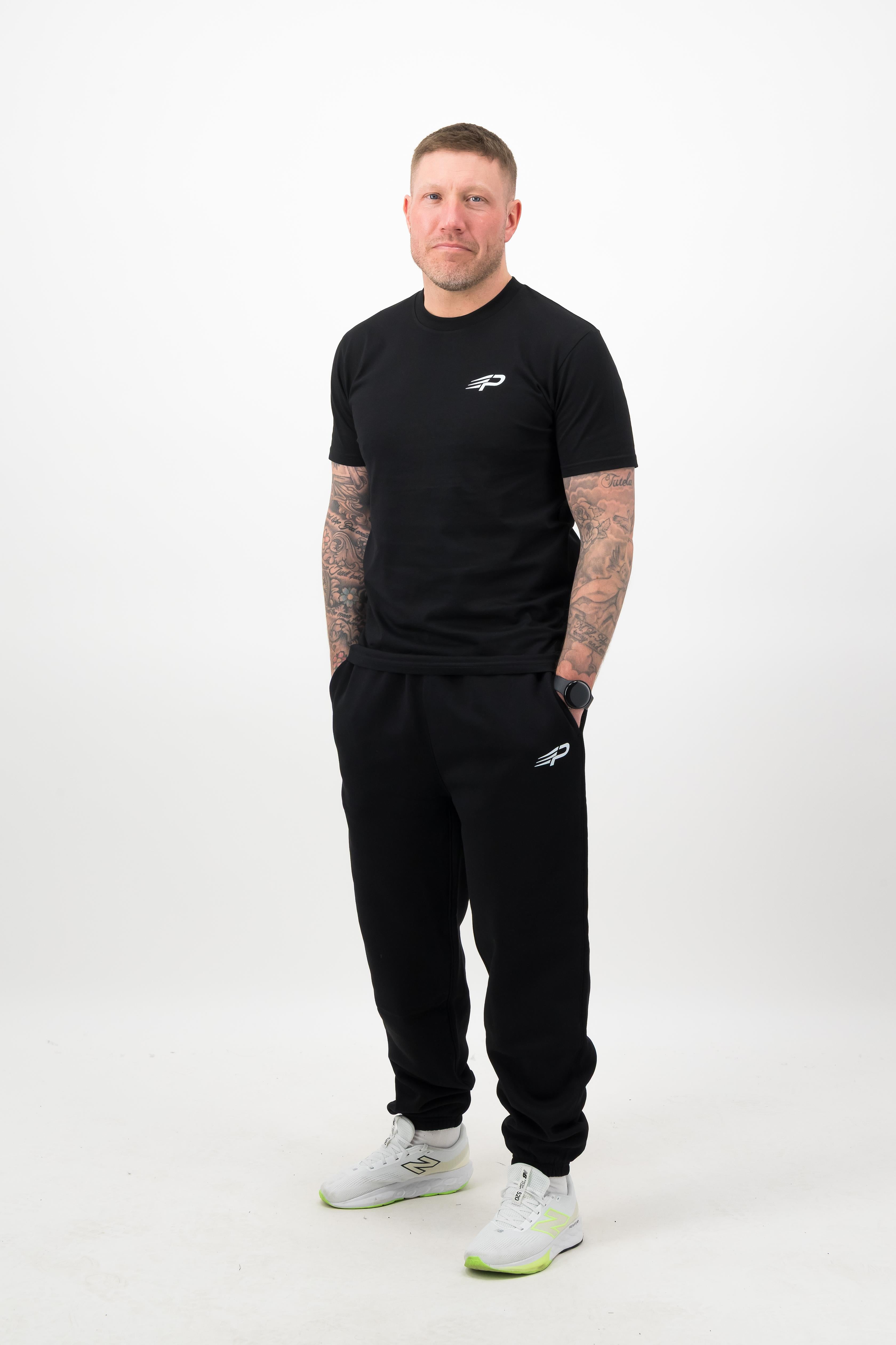 Person wearing a Concept Padel black rebound tee standing facing forward, styled with black joggers and trainers against a white background