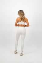 Woman wearing a Concept Padel ecru performance bra standing with back to camera showing the streamlined back silhouette, styled with white leggings and trainers against a white background