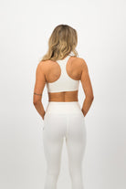 Woman wearing a Concept Padel ecru performance bra standing with back to camera showing the streamlined back silhouette, styled with white leggings and trainers against a white background