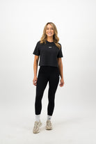 Woman wearing a Concept Padel black faded crop tee standing facing forward, styled with black leggings and trainers against a white background