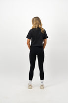 Woman wearing a Concept Padel black faded crop tee standing with back to camera showing the clean back silhouette, styled with black leggings and trainers against a white background