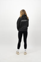 Woman wearing a Concept Padel black faded hoodie standing with back to camera showing the back Concept print, styled with black leggings and trainers against a white background