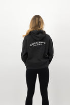 Woman wearing a Concept Padel black faded hoodie standing with back to camera showing the back Concept print, styled with black leggings and trainers against a white background