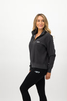 Woman wearing a Concept Padel black faded 1/4 zip sweatshirt standing side on, paired with black leggings and trainers against a white background