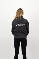 Woman wearing a Concept Padel black faded 1/4 zip sweatshirt standing with back to camera showing the back arch logo, styled with black leggings and trainers against a white background