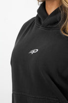 Close‑up of the embroidered Concept Padel logo on the black faded hoodie worn by a woman against a white background
