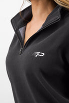Close‑up of the embroidered Concept Padel logo on the black faded 1/4 zip sweatshirt worn by a woman against a white background