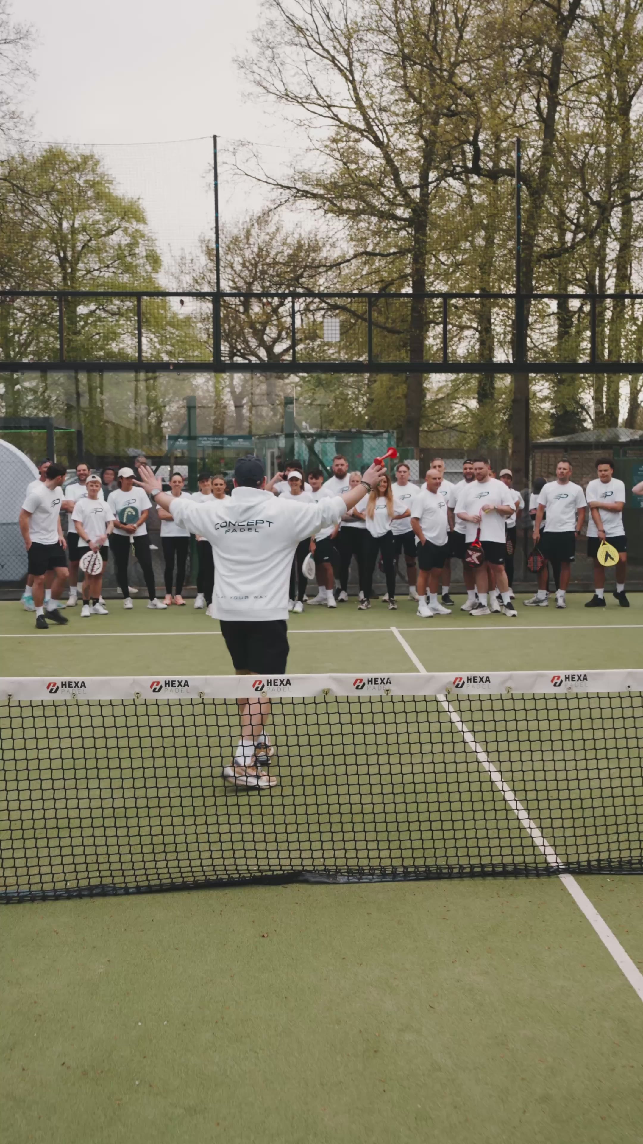 Group of people on an outdoor padel court with one person in a white Concept Padel T‑shirt raising their arms toward the group across the net